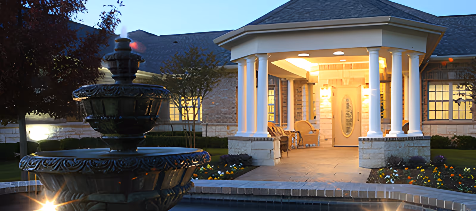 Evening view of the entrance to a senior living facility with a covered porch supported by white columns, warm lighting, and a decorative water fountain in the foreground surrounded by landscaping and flowers.