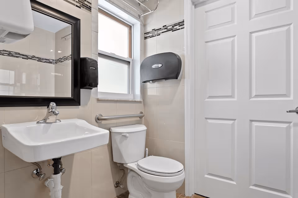 A clean and modern bathroom featuring a white sink with a chrome faucet, a large rectangular mirror with a dark frame, a white toilet with a safety grab bar beside it, a window with frosted glass, and wall-mounted black soap and paper towel dispensers. The walls are tiled with beige tiles and a decorative horizontal strip of smaller tiles near the ceiling.