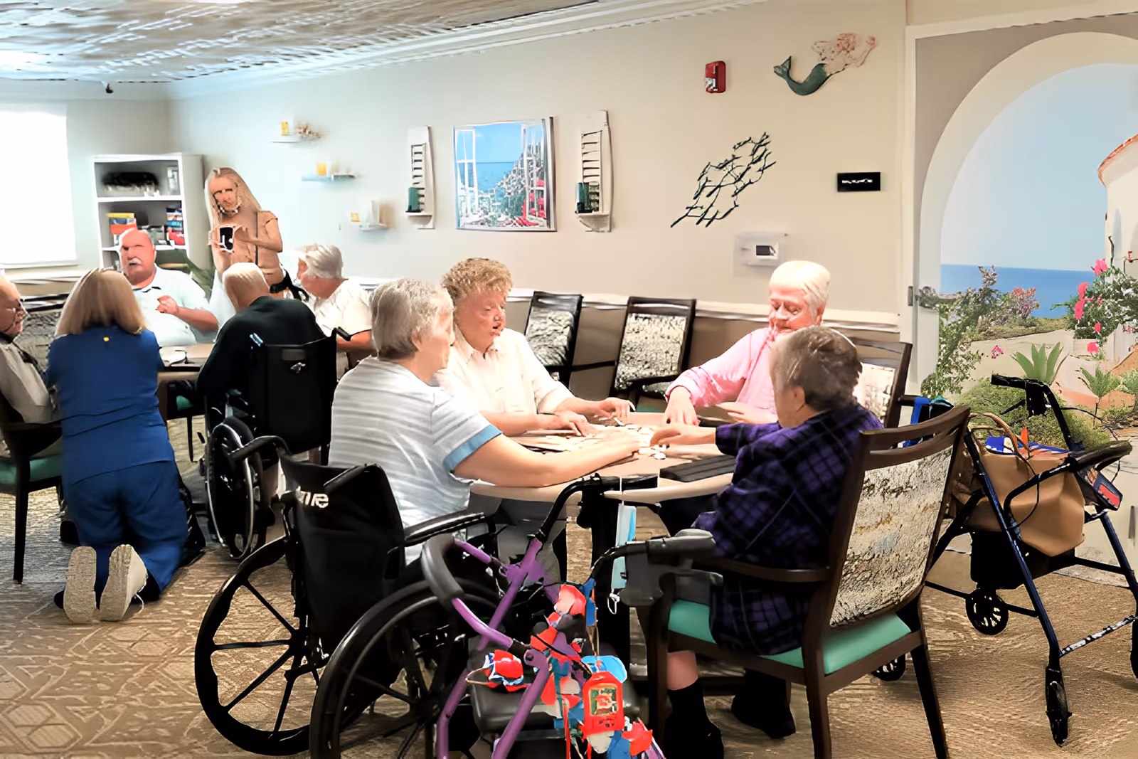 A group of elderly people sitting around tables in a common room, some in wheelchairs, engaging in activities and socializing. A caregiver is kneeling and assisting one of the residents. The room has light-colored walls with decorative wall art and a mural of a seaside scene visible through an archway.