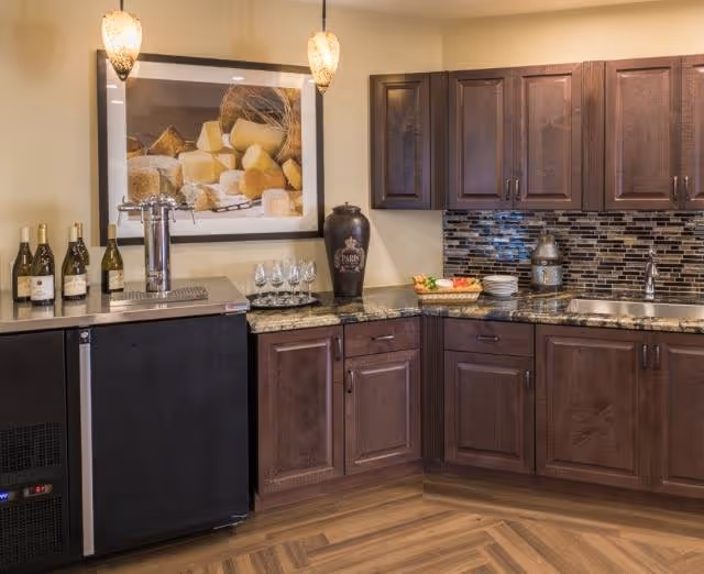 A kitchen area with dark wooden cabinets, a granite countertop, a stainless steel sink, and a tiled backsplash. There are four wine bottles and four wine glasses on the counter, a large decorative vase, a basket with vegetables, and a framed picture of cheese on the wall. Two pendant lights hang from the ceiling.