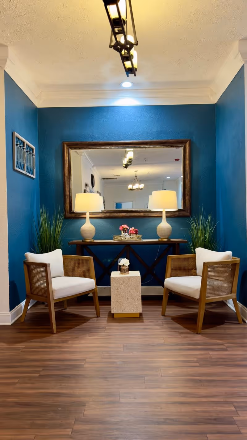 A cozy seating area with two wooden chairs with white cushions facing each other, separated by a small square table with a flower vase on it. Behind the chairs is a wooden console table with two lamps and a decorative tray with flowers. The wall behind the console table is painted blue and features a large rectangular mirror reflecting the ceiling lights and part of the room.