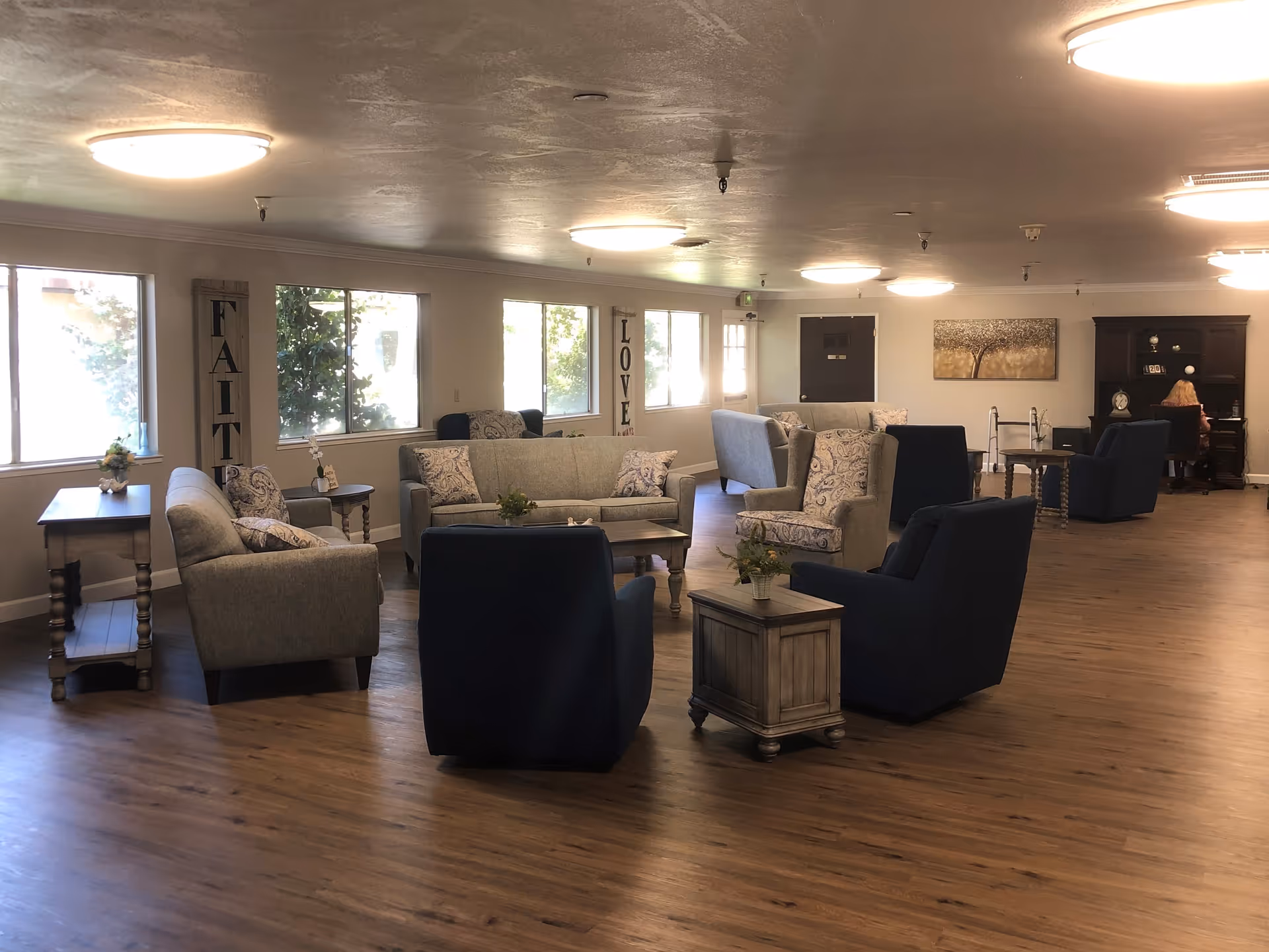 A spacious living room area with multiple seating arrangements including gray sofas, patterned armchairs, and dark blue chairs. The room has large windows letting in natural light, wooden flooring, and soft ceiling lights. There are decorative signs on the walls with the words 'FAITH' and 'LOVE'. In the background, a person is seated at a dark wooden desk with a hutch.