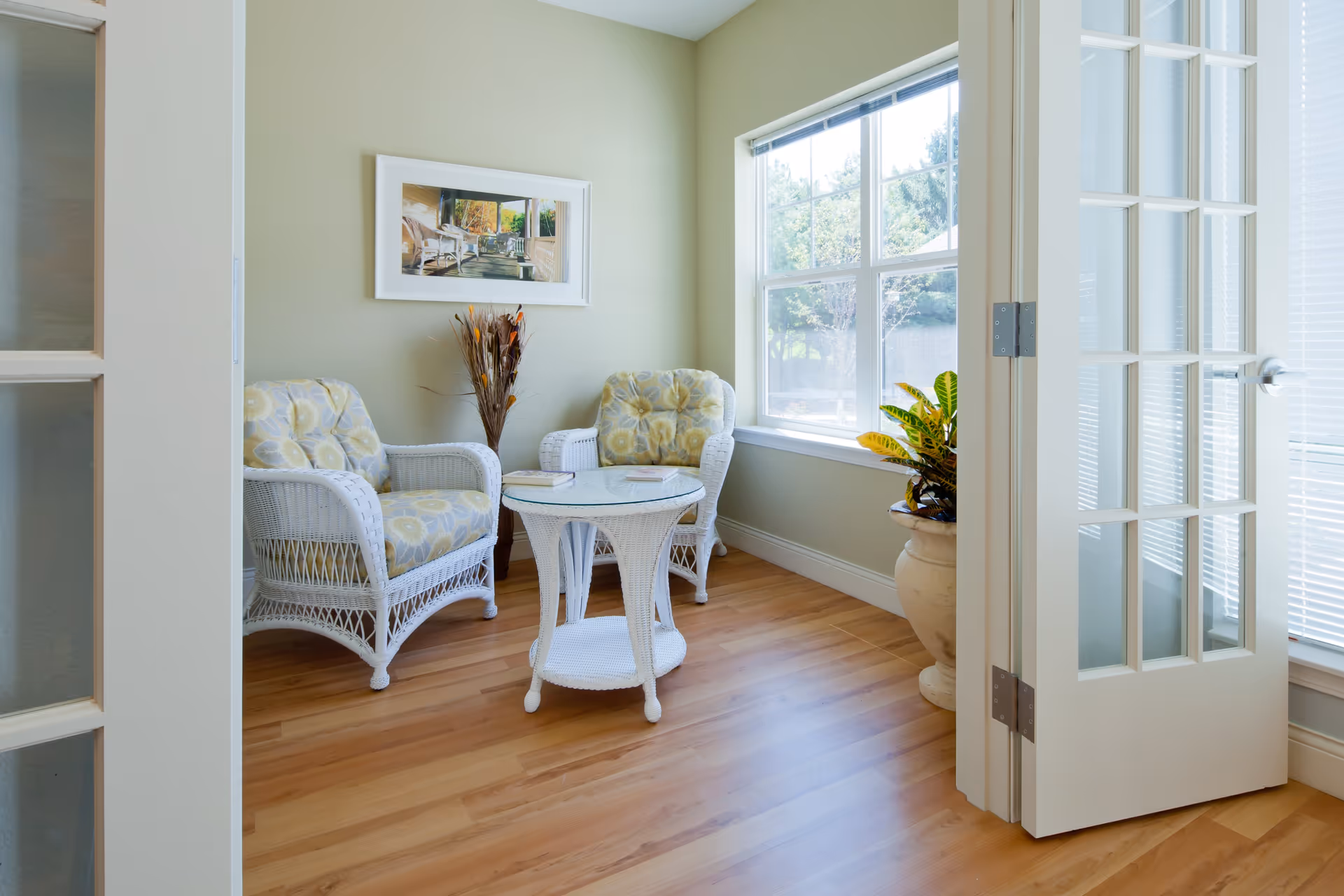 A bright and cozy sitting area with two white wicker armchairs featuring floral cushions, a matching round wicker table with a glass top, a large window letting in natural light, a potted plant in a beige pot, and a framed picture on the light green wall. The room has wooden flooring and white French doors.