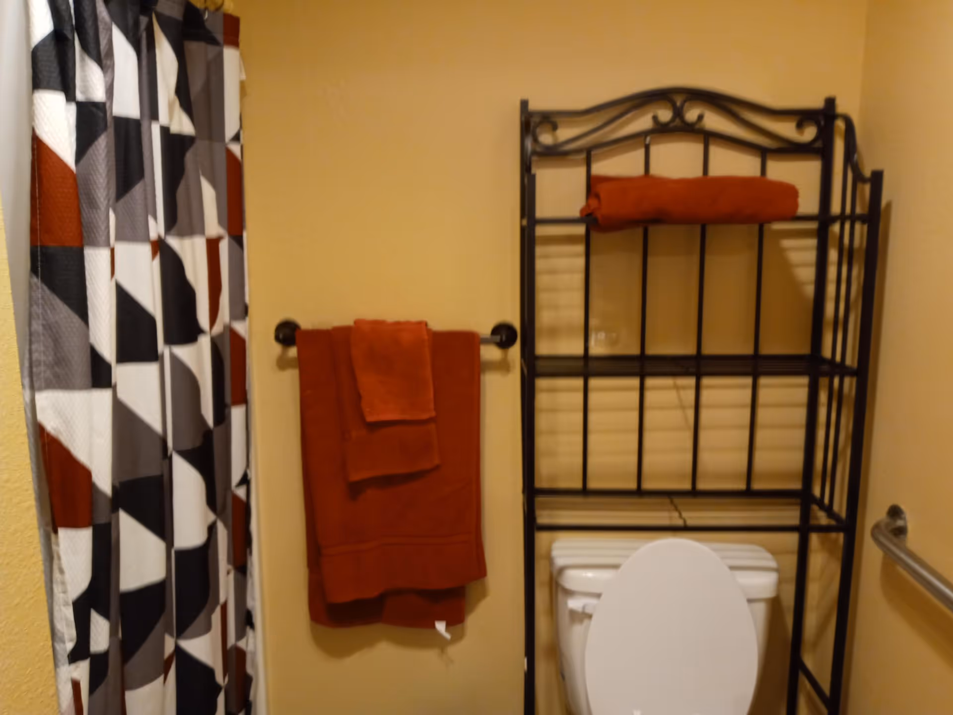 Bathroom with a white toilet, a black metal shelving unit above the toilet holding a folded red towel, a towel rack on the wall with two red towels, and a shower curtain with a geometric pattern in black, white, gray, and red colors.