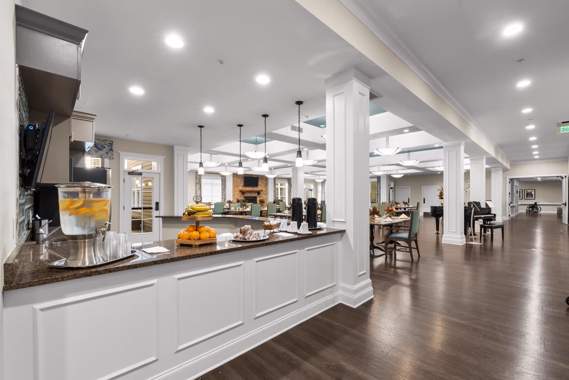 A spacious and well-lit dining area in a senior living facility with a counter in the foreground holding a water dispenser with lemon slices, a tiered fruit tray with bananas and oranges, coffee dispensers, and pastries. The dining room features multiple tables and chairs, a stone fireplace with a TV mounted above it, pendant and ceiling lights, and a piano near the hallway.
