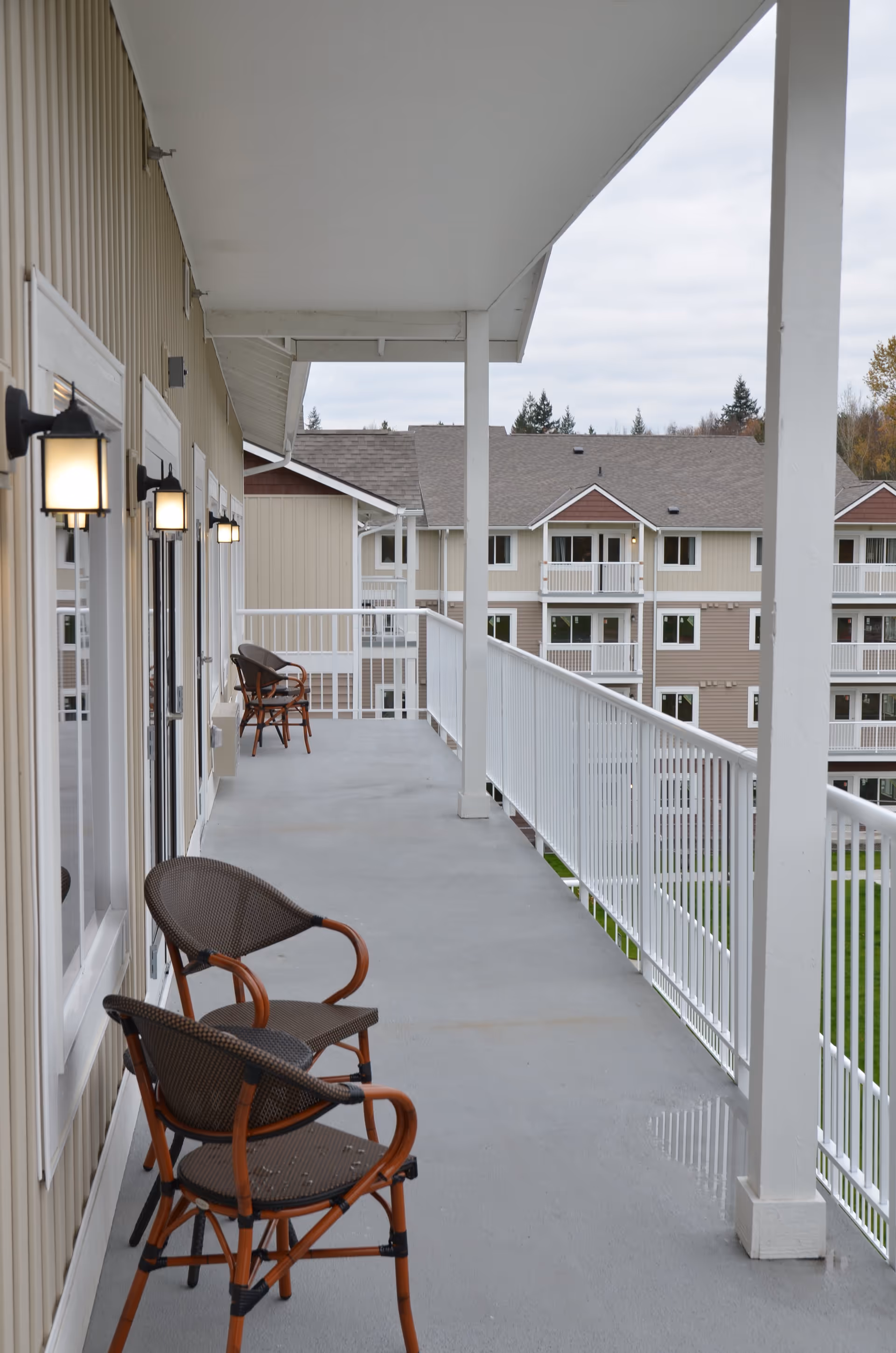 A covered outdoor balcony with a gray floor and white railing, featuring several brown wicker chairs with wooden frames. The balcony overlooks a courtyard with multi-story beige and brown residential buildings in the background under a cloudy sky.