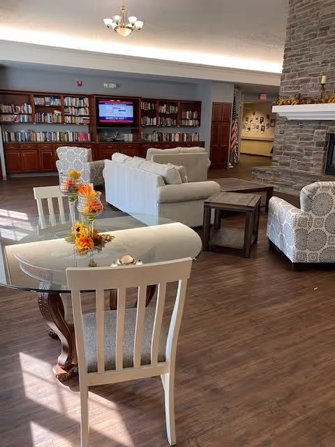A cozy senior living facility common area with a glass-top dining table and white chairs in the foreground, decorated with a fall-themed centerpiece. In the background, there are comfortable upholstered armchairs and sofas arranged around a stone fireplace. A bookshelf filled with books and a mounted TV are visible along the back wall.