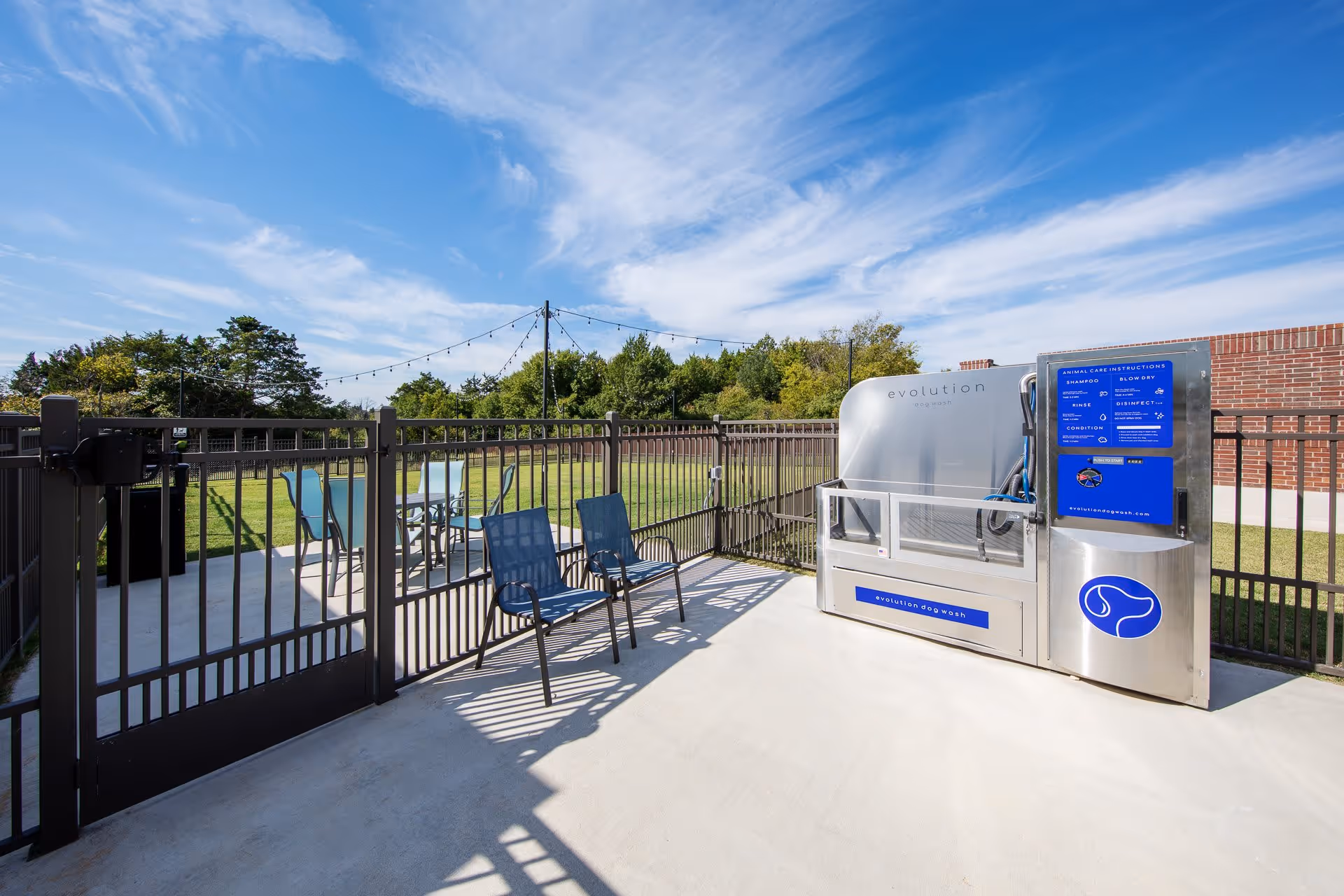 Outdoor fenced area with two blue chairs and a table with chairs in the background, featuring an Evolution dog wash station under a blue sky with scattered clouds.