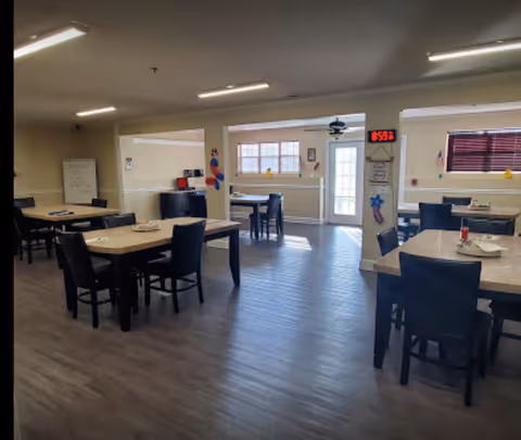 A spacious dining room with several tables and chairs arranged neatly. The room has large windows allowing natural light to enter, a digital clock on the wall showing 8:59, and some decorations on the walls. The floor is wooden, and the ceiling has fluorescent lighting.
