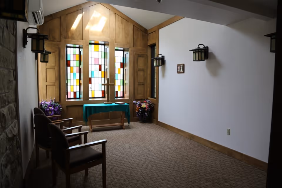 Small chapel-style interior with stained glass windows, a wooden cross on a table, chairs, and wall sconces.