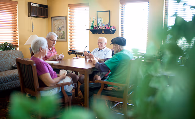 Four elderly people sitting around a wooden table playing cards in a well-lit room with large windows and wooden blinds. The room has a cozy atmosphere with a patterned carpet, a couch, framed artwork on the wall, and a shelf with flowers and a mirror.