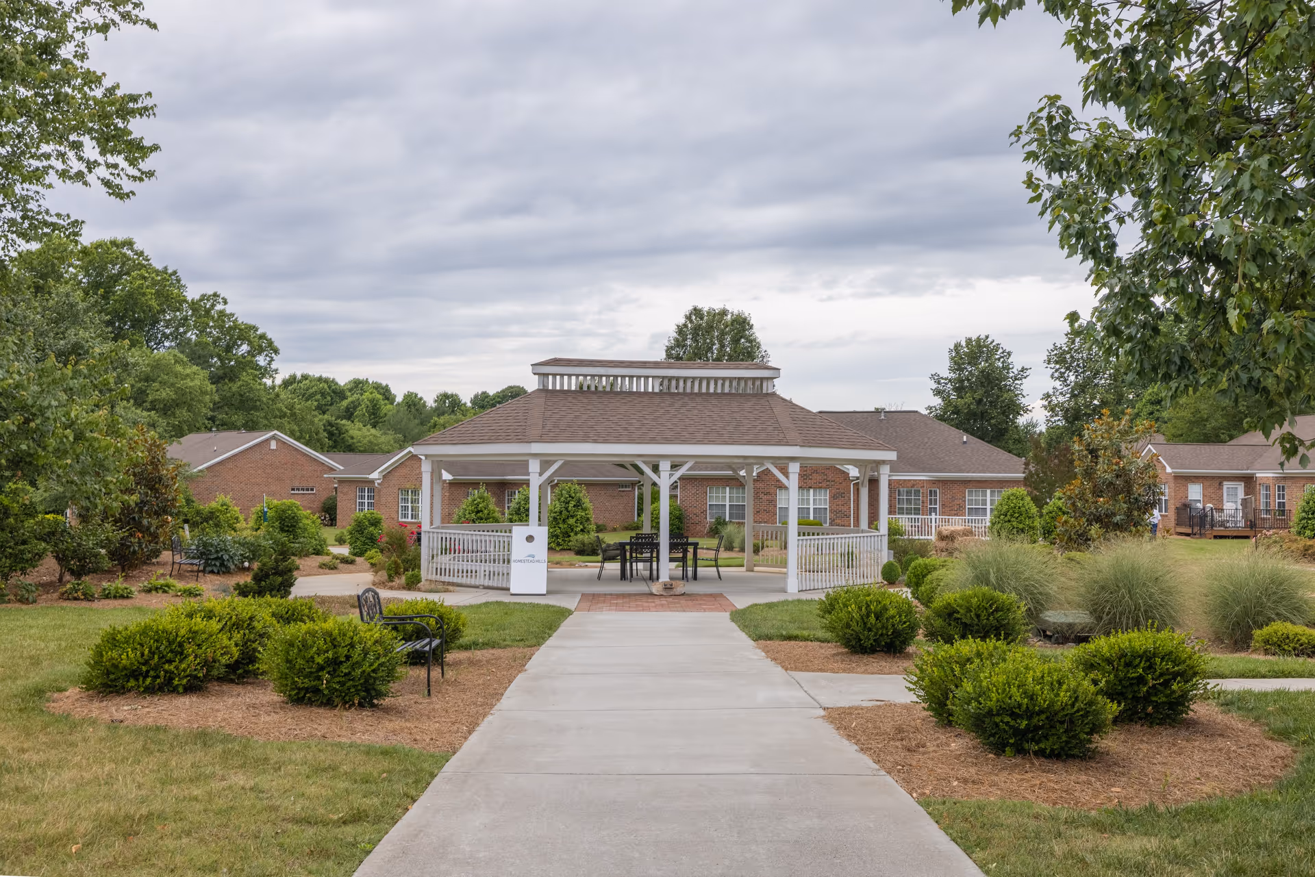 Outdoor view of a senior living facility with a paved walkway leading to a white gazebo surrounded by green bushes and trees. Brick buildings are visible in the background under a cloudy sky.