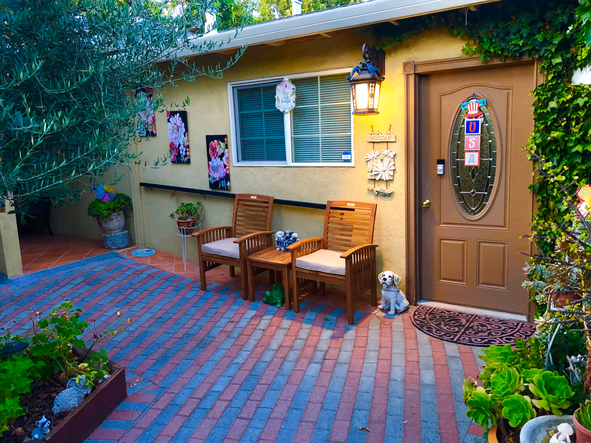 A small courtyard in front of a residence with two wooden chairs, potted plants, wall art, and a brown front door with an oval window and a dog statue.