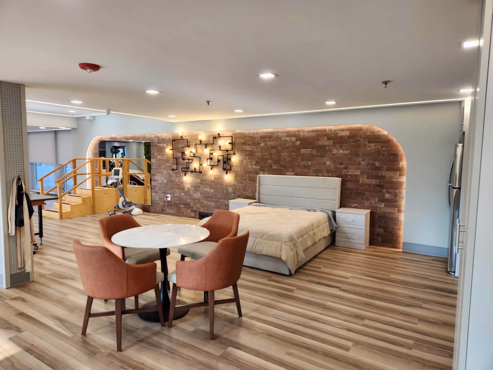 Open studio-style bedroom in a senior living facility with a bed against a lit brick accent wall, a round table with four chairs, and a small exercise area.
