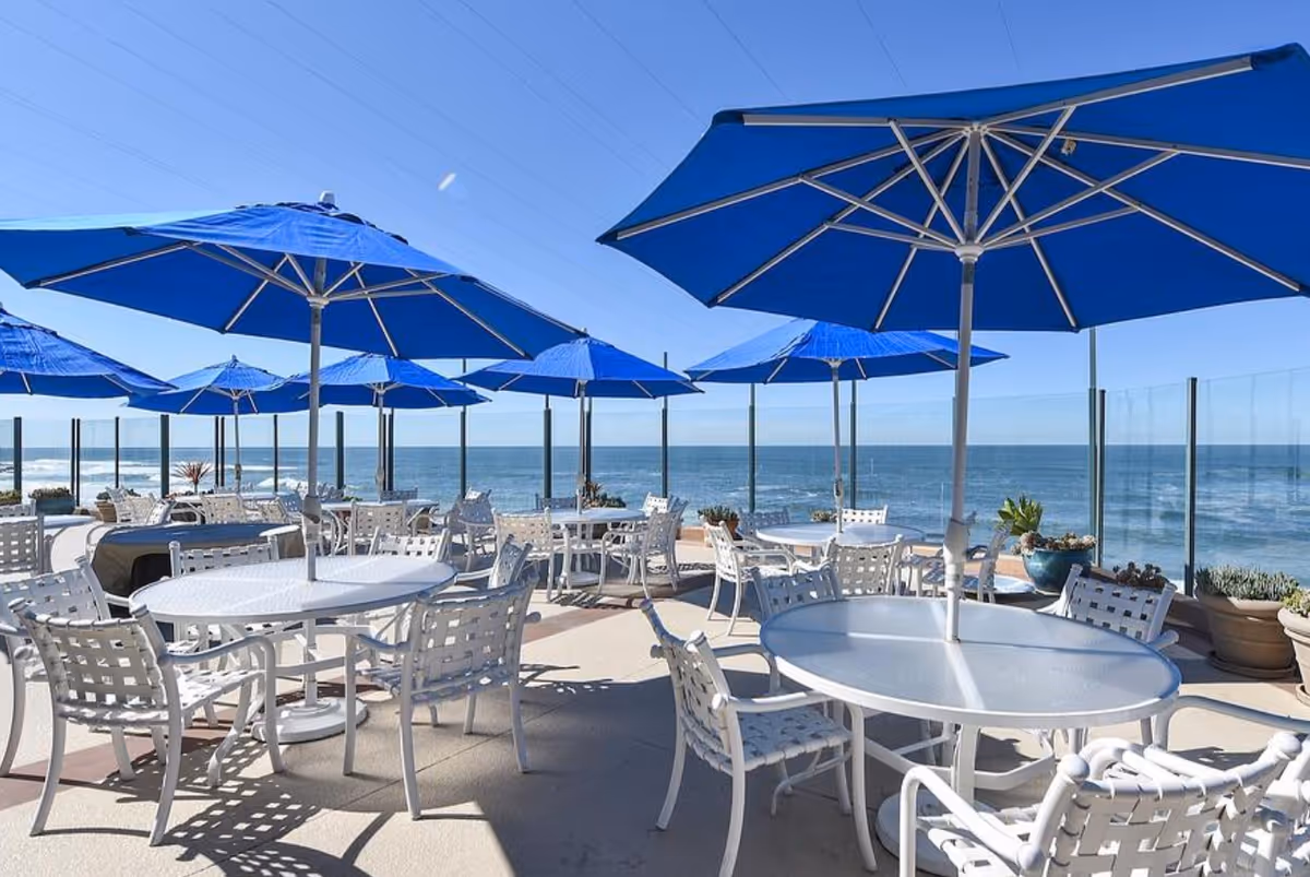 Oceanfront outdoor patio with white tables and chairs under blue umbrellas overlooking the sea.