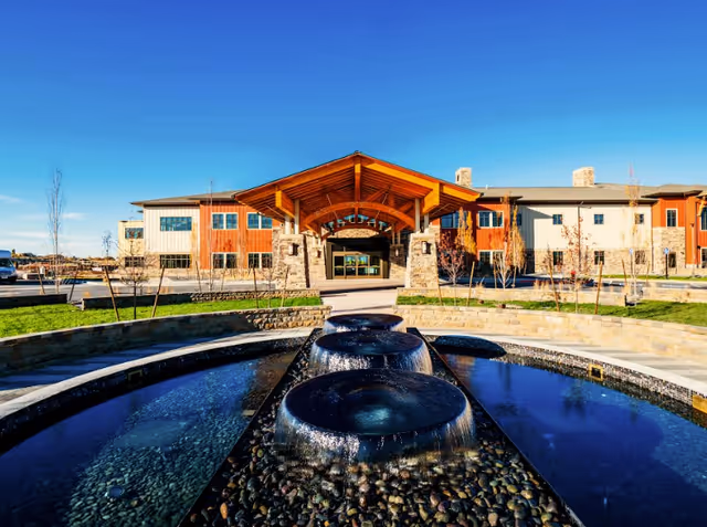 Exterior view of Summit Vista facility with a modern building featuring stone and wood accents, a large covered entrance, and a decorative water fountain in the foreground under a clear blue sky.