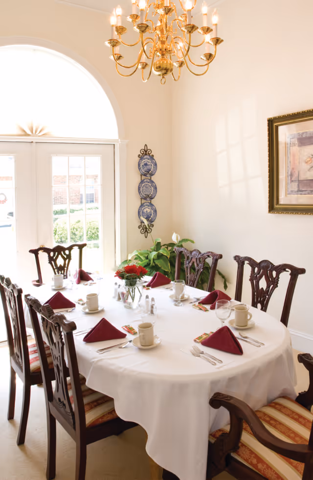 A formal dining room with a round table covered with a white tablecloth, set with cups, glasses, silverware, and folded burgundy napkins. The room features wooden chairs with patterned cushions, a gold chandelier hanging above, a framed artwork on the wall, decorative blue plates on a wall-mounted rack, and a large window with an arched top letting in natural light.