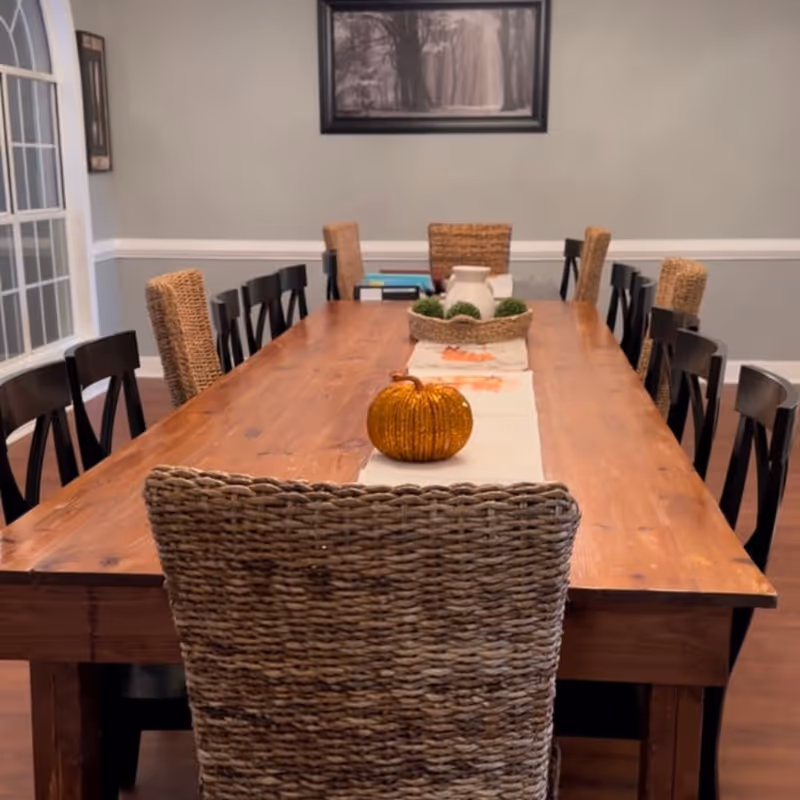 A long wooden dining table with a white runner decorated with a small orange pumpkin and a basket centerpiece containing a white pitcher and green plants. The table is surrounded by a mix of woven and black wooden chairs. The room has light gray walls with white trim, a large window on the left, and a framed black and white forest photograph on the wall.