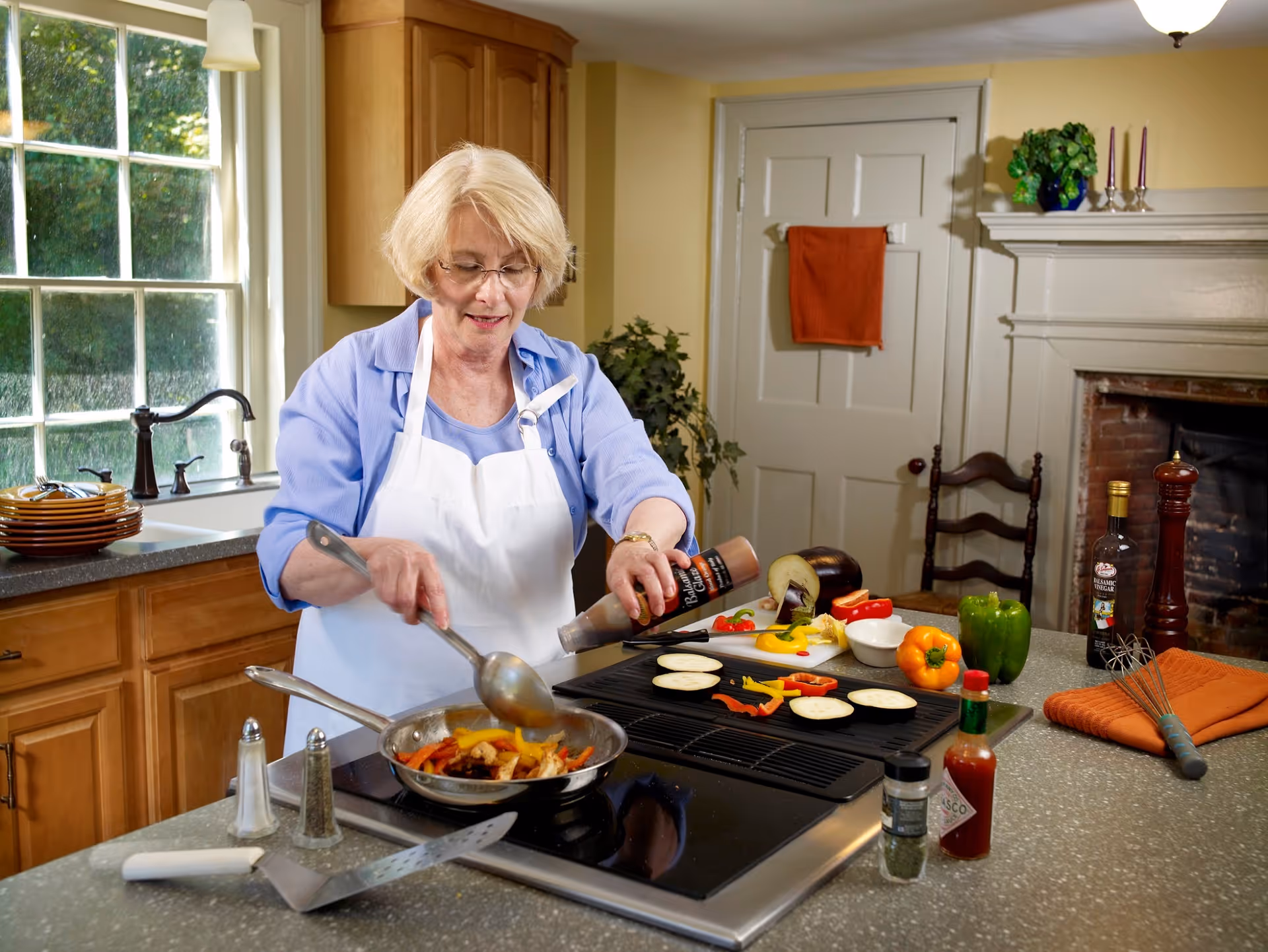 An elderly woman wearing glasses and a white apron is cooking in a kitchen. She is stirring vegetables in a frying pan on a stovetop while pouring sauce from a bottle. The kitchen has wooden cabinets, a window with a view of greenery outside, and various vegetables and condiments on the countertop.