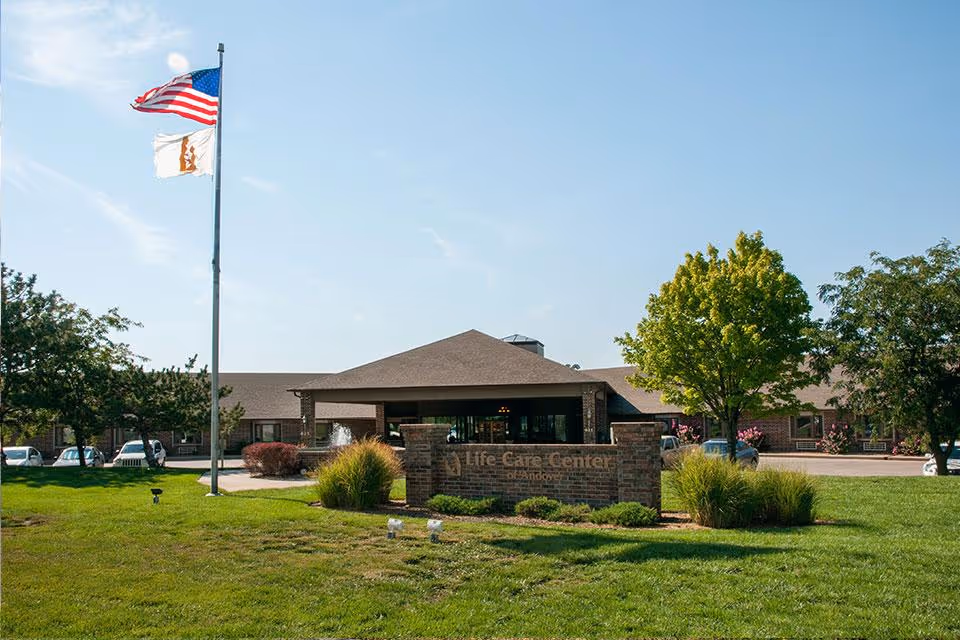Front exterior view of Life Care Center of Andover building with a brick sign in front, green lawn, trees, and two flagpoles with the American flag and another flag against a clear blue sky.