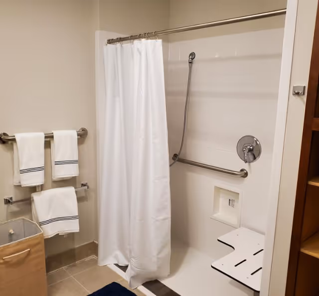 A bathroom shower area with a white shower curtain, a fold-down shower seat, and a handheld showerhead. There are grab bars installed on the walls for safety. Three white towels with black stripes hang on towel racks next to the shower. A laundry hamper is visible on the floor, and part of a wooden shelving unit is seen on the right side.