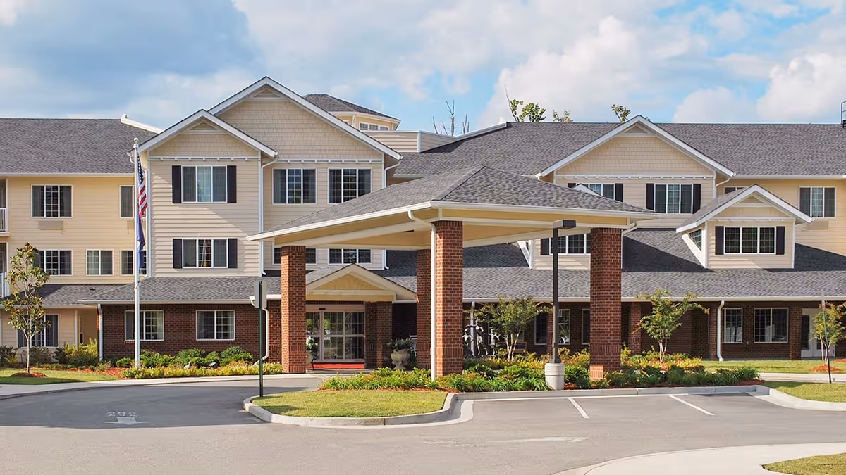 Front exterior view of a senior living facility named Dogwood Terrace by Barclay House, featuring a large covered entrance supported by brick columns, multiple windows, and a well-maintained garden area with small trees and shrubs. The sky is partly cloudy.