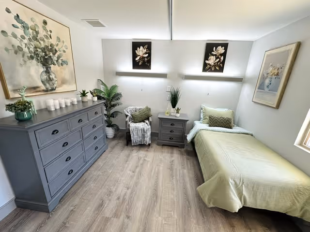 A neatly arranged bedroom with a single bed covered in a light green bedspread, a gray nightstand with decorative plants, a gray dresser with candles and a small plant, a cozy chair with a throw blanket, and framed floral artwork on the walls. The room has light wood flooring and soft lighting.