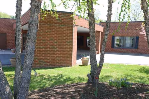 Brick senior living facility entrance with trees, lawn, and planters along a covered walkway.