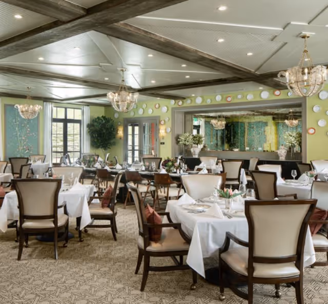 Elegant dining room with round tables set with white linens, upholstered chairs, chandeliers, and decorative plates on green walls.
