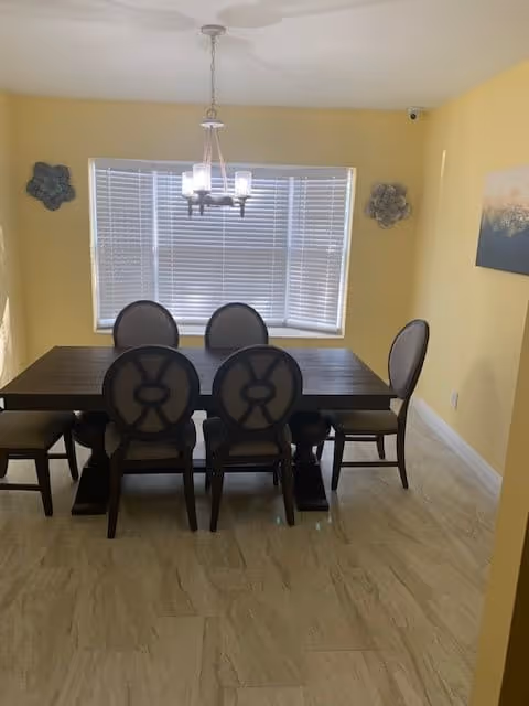 A dining room with a dark wooden rectangular table surrounded by six chairs with circular backrests. The walls are painted yellow, and there is a window with white blinds behind the table. A chandelier with five lights hangs above the table. Decorative wall art pieces are mounted on either side of the window, and a painting is hung on the right wall.