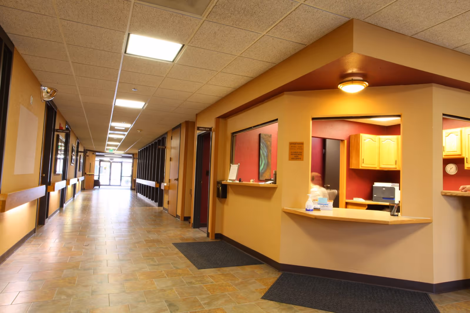 Interior hallway of a senior living facility with a reception desk on the right side, beige walls, tiled floor, and ceiling lights. The hallway extends to a glass exit door at the far end.