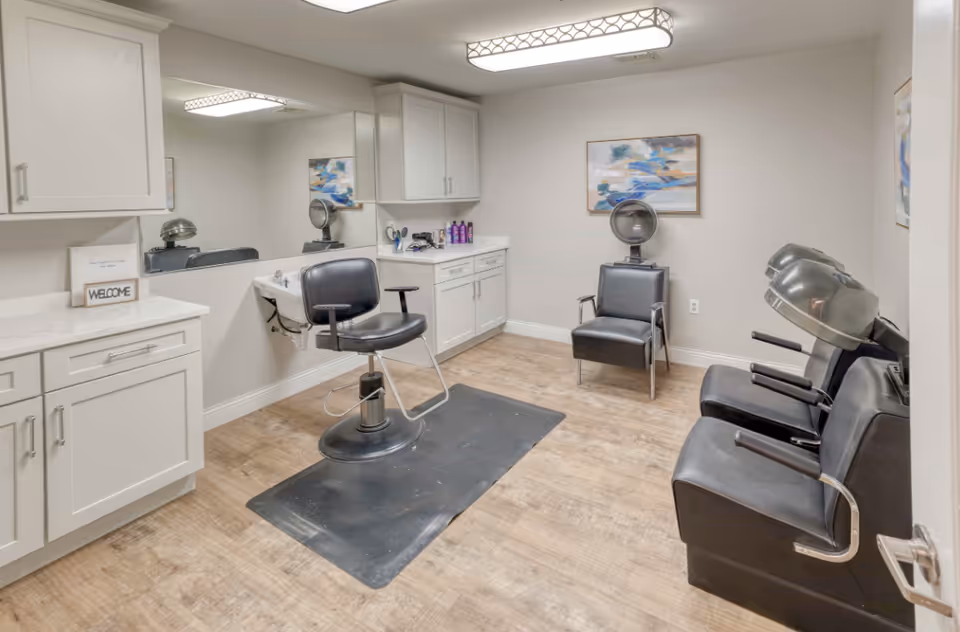Interior view of a hair salon area in a senior living facility with a black salon chair in front of a sink and mirror, two black hair drying chairs with hooded dryers, white cabinets, wooden flooring, and a colorful abstract painting on the wall.