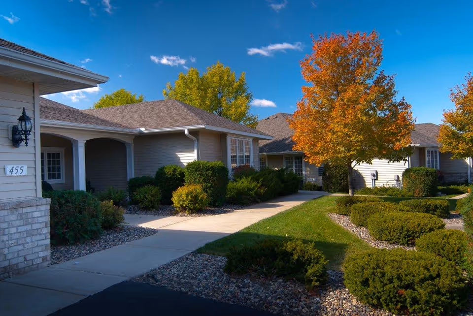 Exterior view of a senior living facility with beige buildings, well-maintained landscaping including bushes and trees with autumn foliage, a clear blue sky, and a sidewalk leading through the grounds.