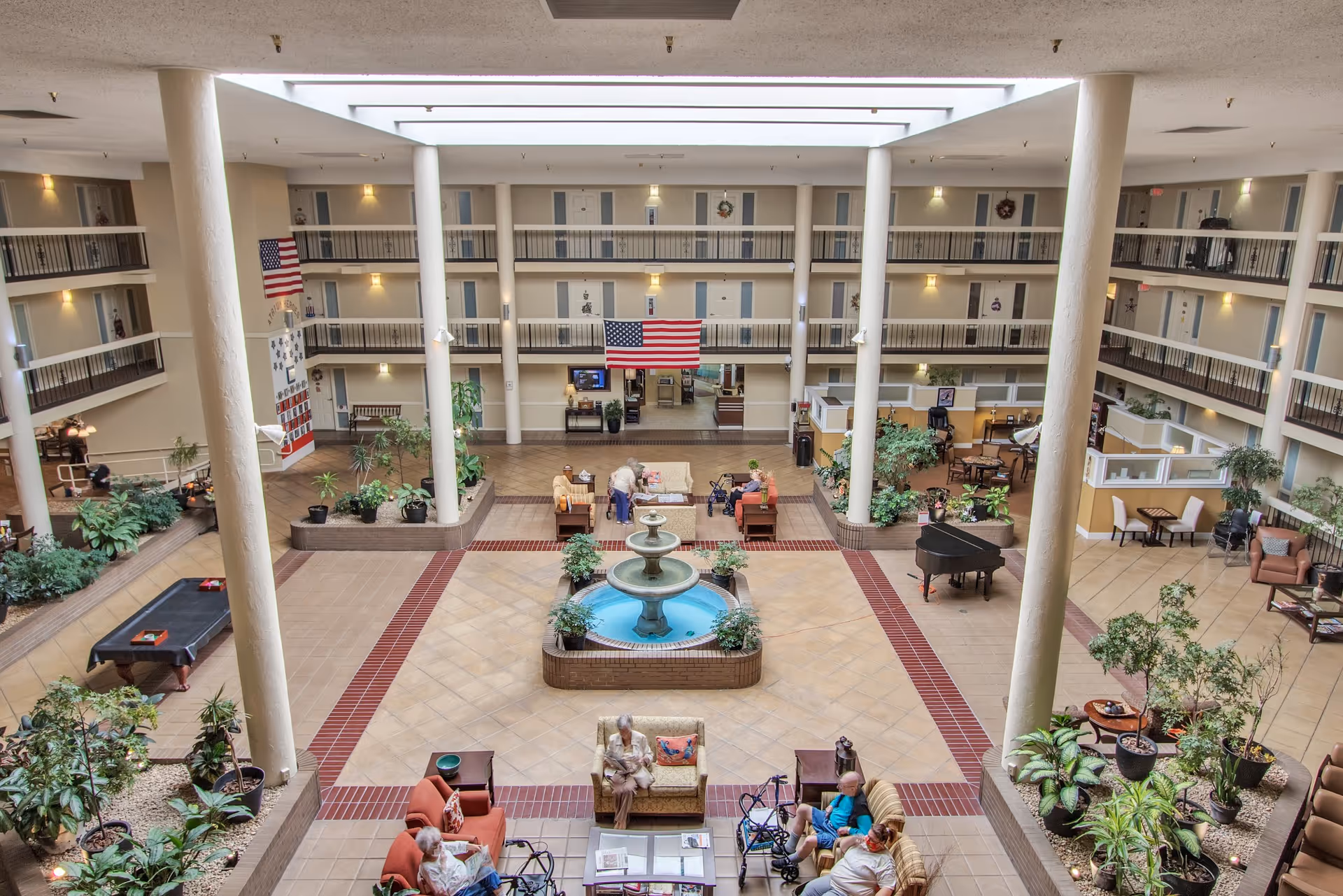 Multi-story atrium of a senior living facility with a central fountain, seating areas, plants, and balconies overlooking the space.