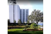 A tall white multi-story building with numerous windows, surrounded by trees and greenery, with a well-maintained lawn and garden beds in the foreground under a clear sky.