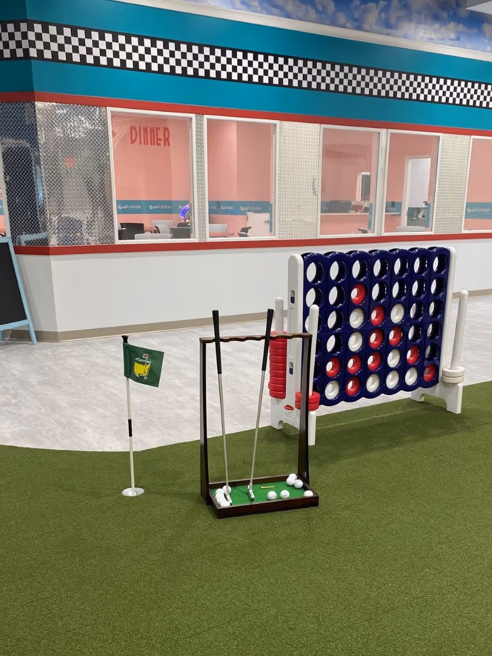 Indoor recreational area with artificial green turf featuring a mini golf setup including a small putting green, golf clubs, and balls. Nearby is a large Connect Four game with red and white discs. The background shows a diner-themed room with pink walls, windows, and a checkered black and white border near the ceiling.