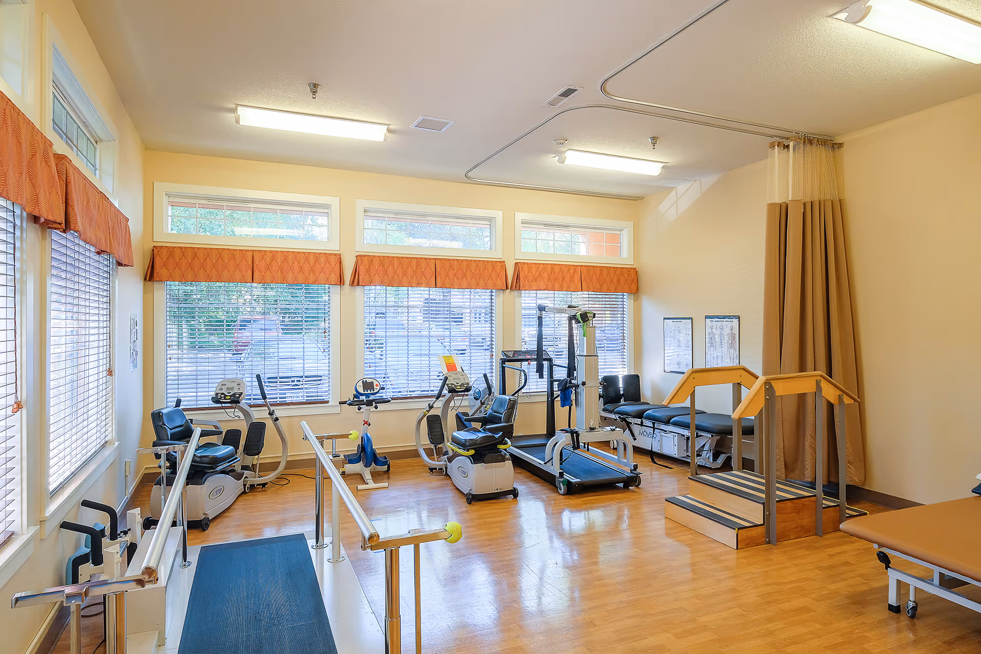 Bright rehabilitation gym with exercise bikes, a treadmill, parallel bars and other therapy equipment in a room with large windows.