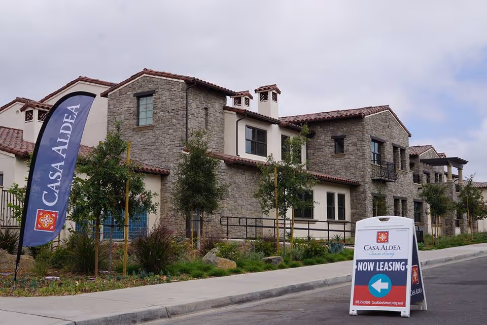 Stone-clad senior living building with Casa Aldea flags and a 'Now Leasing' sidewalk sign in front.
