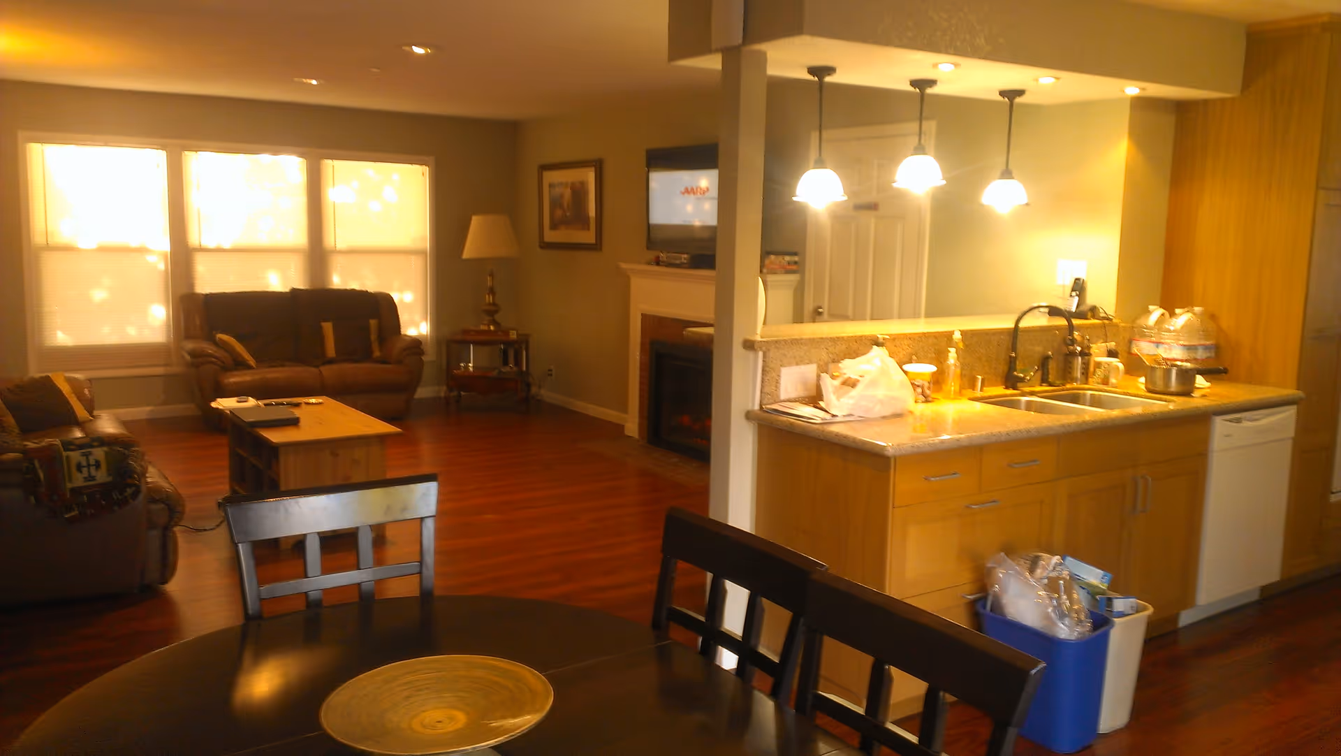 Open-plan living area with a kitchen island and sink under pendant lights, a dining table in the foreground, and sofas by sunlit windows.