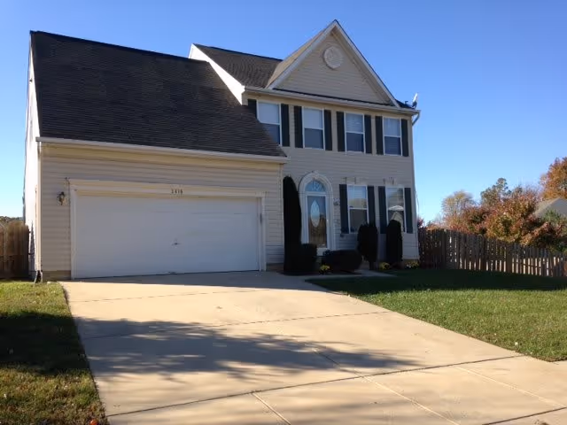 A two-story beige house with a double garage, a concrete driveway, green lawn, and a wooden fence on the right side under a clear blue sky.