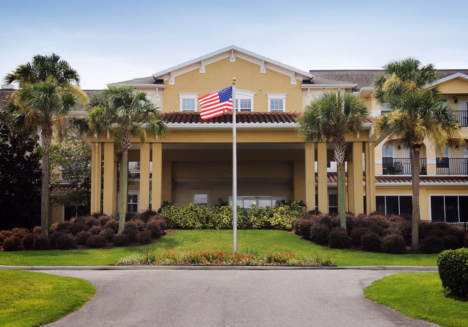 Front exterior view of a yellow senior living facility building with a covered entrance, palm trees, and an American flag on a flagpole in the center of a landscaped area.