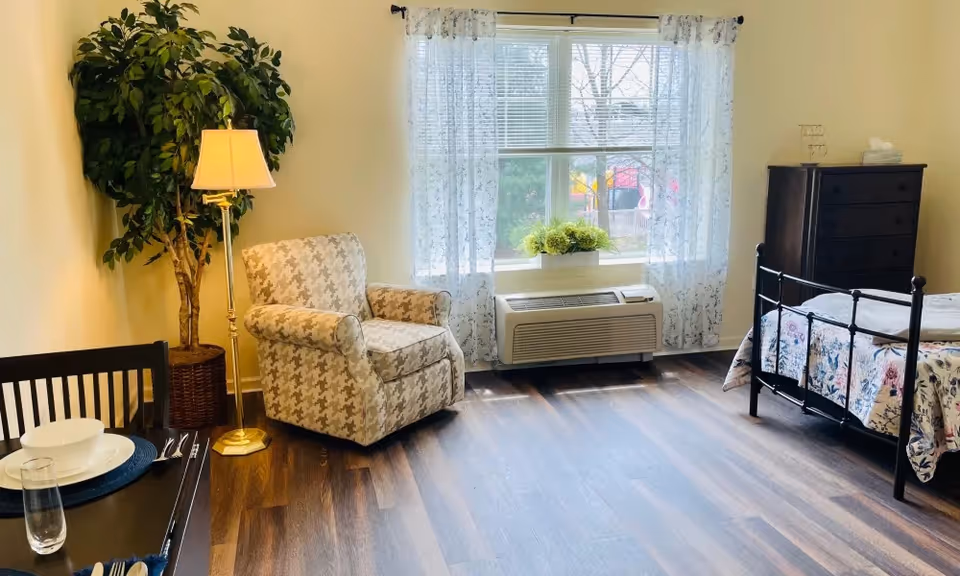 Sunny studio bedroom with an upholstered armchair and floor lamp, a metal-framed bed with floral bedding, a dresser, and a window with sheer curtains above an AC unit.
