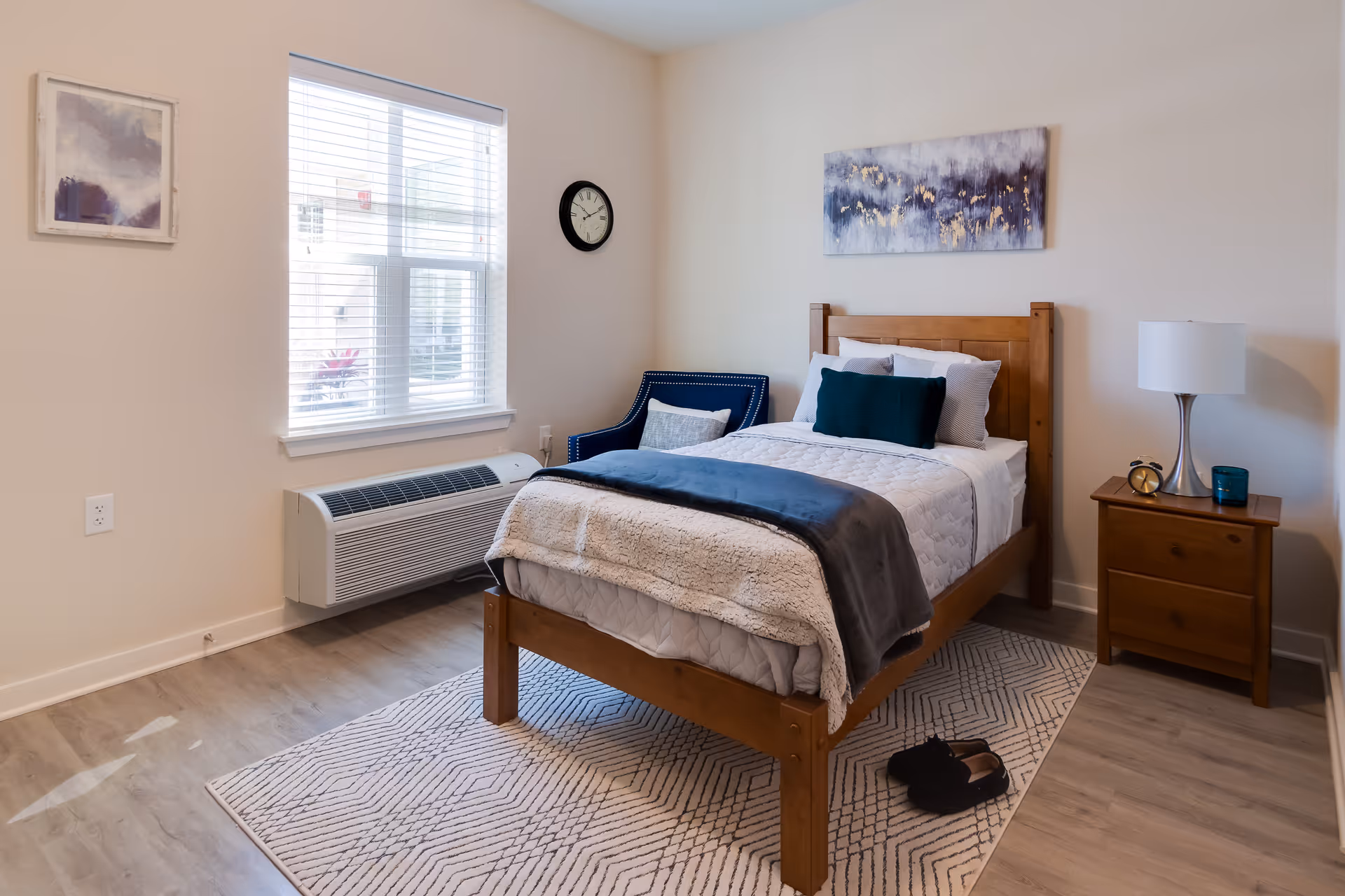 A cozy bedroom with a wooden single bed made with white and gray bedding, a dark blue throw blanket, and decorative pillows. Next to the bed is a wooden nightstand with a lamp, an alarm clock, and a small blue candle. A blue armchair with a pillow is positioned near a window with white blinds. The room has light-colored walls, a wall clock, and two pieces of abstract artwork. A patterned rug is on the wooden floor, and a pair of black slippers is placed beside the bed.
