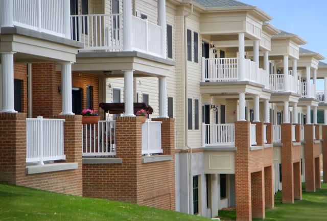 Exterior view of a multi-story senior living building with brick lower levels and rows of covered balconies overlooking a grassy lawn.