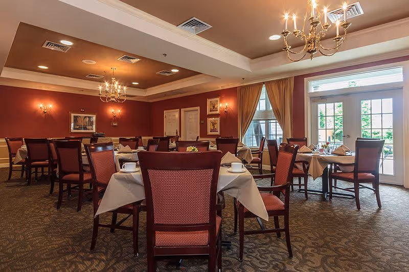 A dining room with multiple tables covered with white tablecloths, set with cups and napkins. The room has red walls, carpeted floors, chandeliers, and large windows with curtains letting in natural light.
