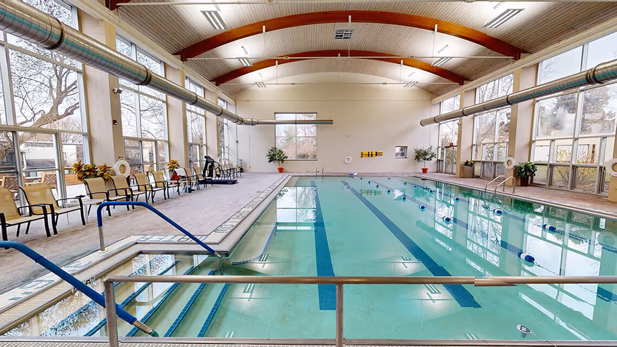 Indoor swimming pool with clear water, lane dividers, and handrails. The pool area has large windows letting in natural light, chairs lined up along the side, potted plants, and a high wooden ceiling with exposed beams.