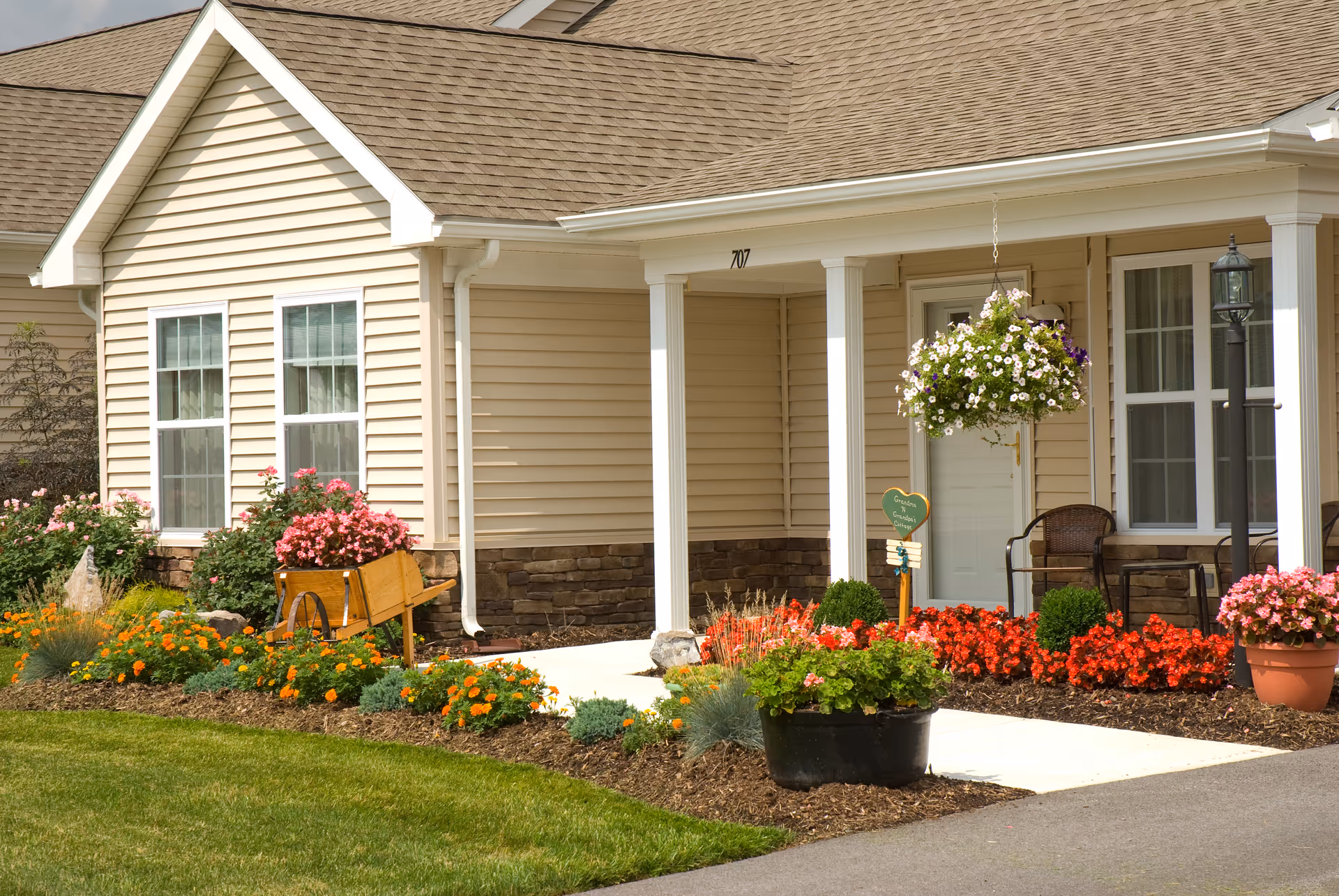 Front porch of a beige siding house with white columns, a hanging flower basket and colorful flower beds lining the walkway.