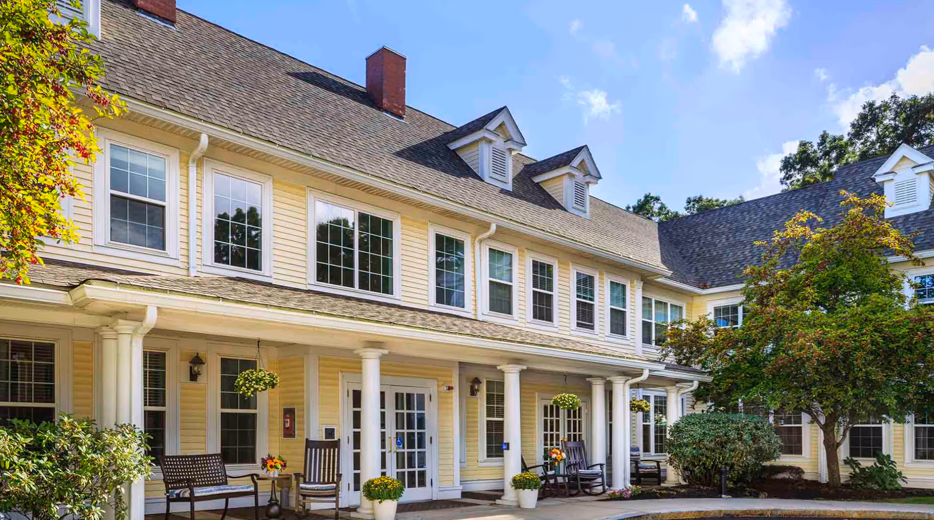 Exterior view of Atria Merrimack Place, a senior living facility with yellow siding, white columns, and multiple windows. The entrance features a covered porch with rocking chairs, hanging flower baskets, potted plants, and surrounding greenery including trees and bushes under a partly cloudy blue sky.