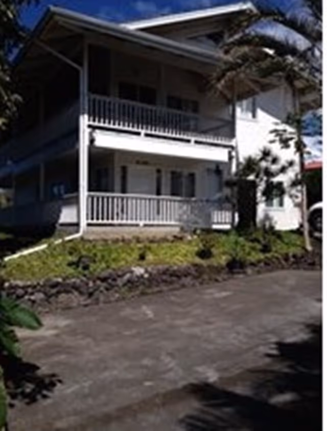 Exterior view of a two-story white building with a covered porch and balcony, surrounded by greenery and palm trees under a blue sky.