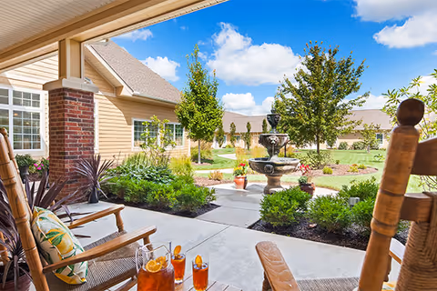 A sunny outdoor patio area at Legend at Capital Ridge featuring wooden rocking chairs with cushions, a small table with glasses of iced tea, a stone fountain surrounded by plants and flowers, and a well-maintained garden with trees and shrubs under a blue sky with scattered clouds.